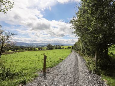 A gravel path running alongside a green field with trees under a cloudy sky at Meadow Lodge in Maerdy near Corwen