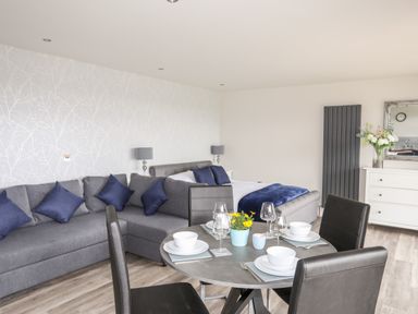 A room with a grey sofa with blue cushions a round dining table set with white bowls and glasses a bed with blue blanket a white dresser with flowers and a mirror at Meadow Lodge in Maerdy near Corwen