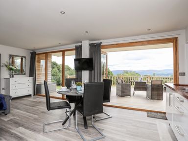 A dining area with a round table and black chairs next to large glass doors opening to a deck with outdoor furniture at Meadow Lodge Maerdy near Corwen