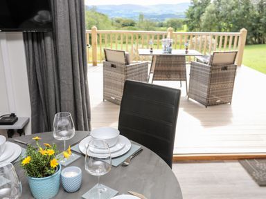 A dining table set with plates bowls glasses and a plant next to a deck with chairs and a table overlooking a green landscape at Meadow Lodge in Maerdy near Corwen
