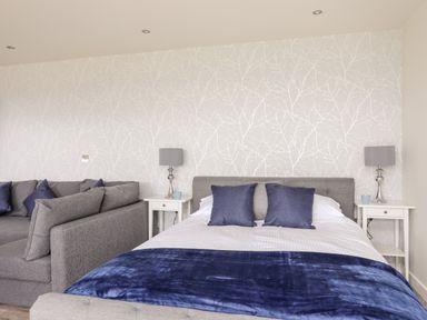 A bedroom with a double bed, gray sofa, two side tables with lamps and patterned wallpaper at Meadow Lodge in Maerdy near Corwen