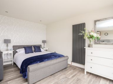 A bedroom with a gray bed, white bedside tables with lamps, a white dresser with flowers and a mirror at Meadow Lodge in Maerdy near Corwen