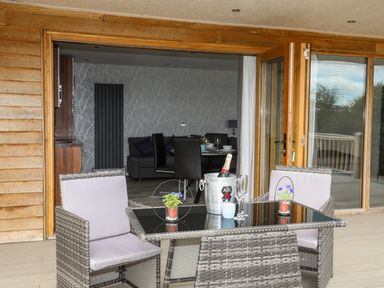 An outdoor patio with a table and chairs set with champagne and glasses next to sliding glass doors opening into a dining area at Meadow Lodge Maerdy near Corwen
