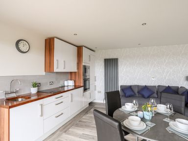 A kitchen with white cabinets and wooden countertop next to a dining table set for four and a gray sofa at Meadow Lodge in Maerdy near Corwen