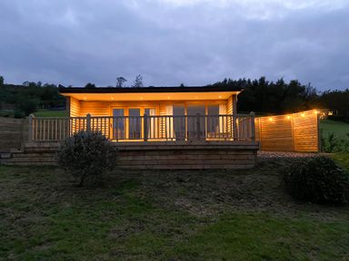 A wooden cabin with lights on a deck and a small tree in front at Meadow Lodge Maerdy near Corwen