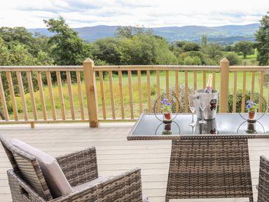 A deck with wicker chairs and a glass table with champagne and flowers overlooking trees and hills at Meadow Lodge in Maerdy near Corwen