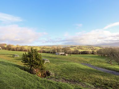 A grassy field with trees picnic benches and hills under a blue sky at Meadow Lodge Maerdy near Corwen