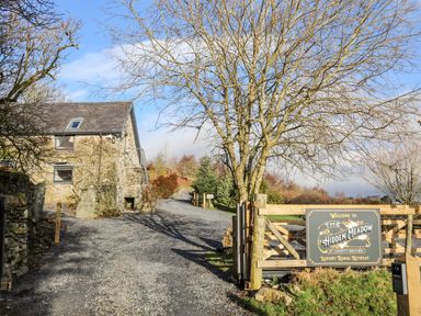 A stone building beside a gravel driveway with a wooden gate and sign for The Hidden Meadow luxury rural retreat