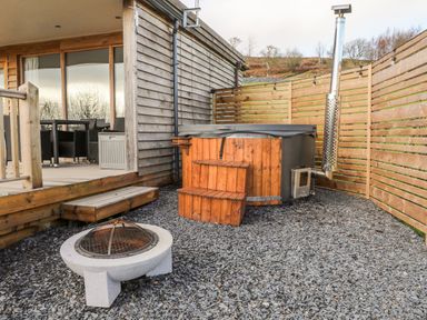 An outdoor area with a wooden hot tub with steps and a fire pit on gravel beside a wooden deck with chairs at Meadow Lodge in Maerdy near Corwen
