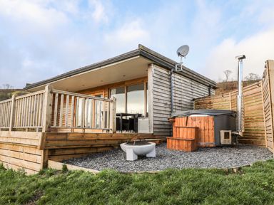 A wooden cabin with a deck railing a fire pit and a hot tub on a gravel area at Meadow Lodge Maerdy near Corwen