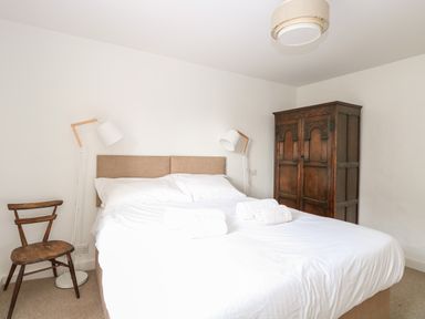 A bedroom with a bed white bedding wooden chair two floor lamps and a wooden wardrobe at Enterprise House in Bury St Edmunds