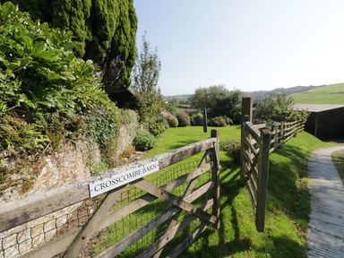 A wooden gate with a crosscombe barn sign opens to a garden with grass and shrubs at Crosscombe Barn in Loddiswell
