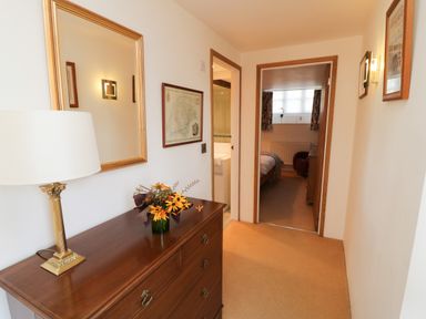 A hallway with a wooden chest of drawers a lamp and flowers on top and doorways leading to a bathroom and bedroom at Crosscombe Barn in Loddiswell
