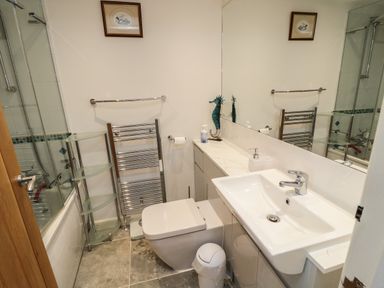 A small bathroom with a toilet sink glass shelves towel rack and a bathtub with shower at Crosscombe Barn in Loddiswell