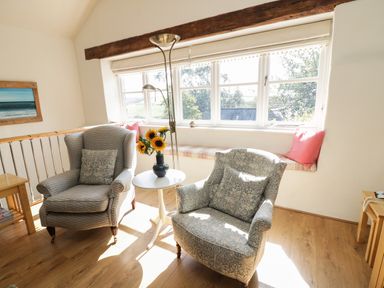 A sitting area with two armchairs a small white table with sunflowers a window seat with cushions and a floor lamp at Crosscombe Barn in Loddiswell