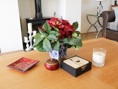 A wooden table with a vase of red flowers green leaves a red dish a small container a set of coasters and a white candle in a living room at Crosscombe Barn in Loddiswell