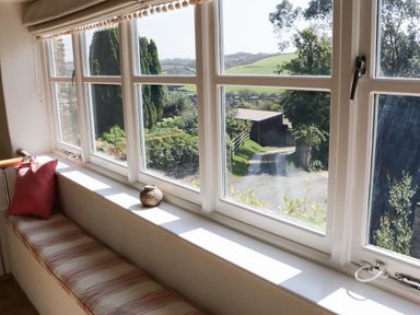 A window seat with a cushion and a decorative item beside large windows overlooking a garden and a driveway at Crosscombe Barn in Loddiswell