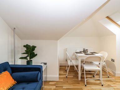 A small dining area with a white table and four white chairs beside a blue sofa with an orange pillow and a plant on a side table at Riviera Heights Paignton