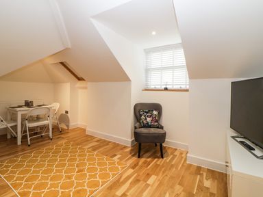 A room with a gray chair with a patterned cushion near a window with blinds a television on a white cabinet a small white dining table with two white folding chairs and a yellow patterned rug on a wooden floor at Riviera Heights in Paignton