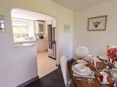 A dining area with a wooden round table set with plates cups and cutlery next to a kitchen with white cabinetry and a window at Greenstyles in Bowness-On-Windermere