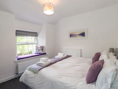 A bedroom with a bed with white sheets and purple pillows and a window seat with a cushion at Greenstyles in Bowness-On-Windermere