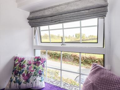 A window with gray blinds two cushions and a view of a road and green fields at Greenstyles in Bowness-On-Windermere