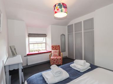 A bedroom with a bed, folded towels, a chair with a cushion, a window seat, a desk, and grey wardrobes at Greenstyles in Bowness-On-Windermere