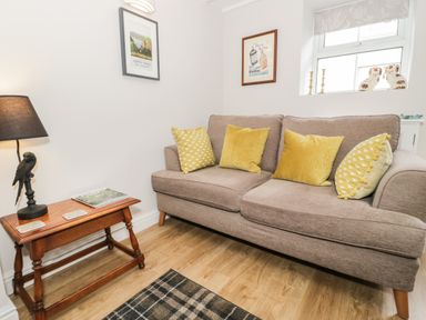 A living room with a beige sofa and yellow cushions next to a wooden side table with a lamp and magazines at Marlin Cottage in Conwy
