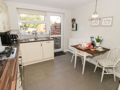 A kitchen with white cabinets a wooden countertop a sink a small dining table with two chairs and a door leading outside at Marlin Cottage in Conwy