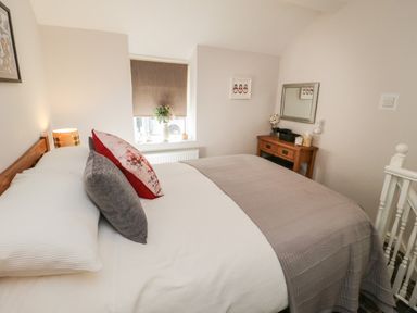 A bedroom with a bed gray and floral pillows next to a window with a table lamp and a wooden desk with a mirror and flowers at Marlin Cottage in Conwy