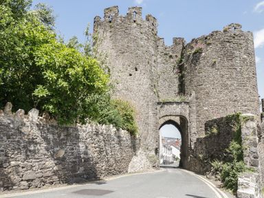 A stone castle gate with two round towers and an arched entrance over a road with trees and stone walls at Marlin Cottage in Conwy