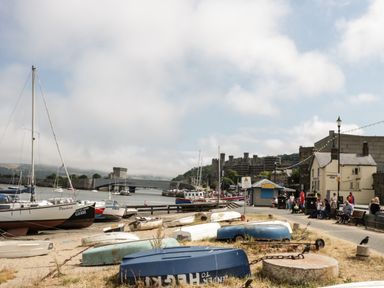 A harbor scene with boats on the shore, a bridge, people sitting on benches, and a castle in the background at Marlin Cottage in Conwy