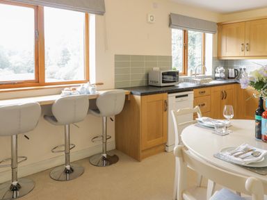 A kitchen with wooden cabinets a counter with a microwave and sink three bar stools by a window and a dining table set with plates and glasses at Hendre in Penybontfawr