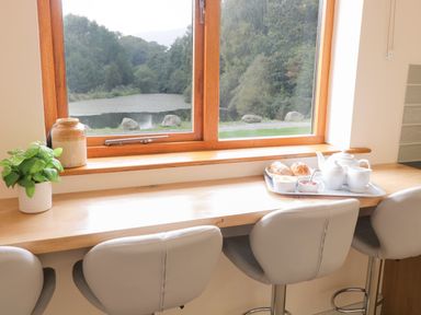 A kitchen counter with gray chairs below a window showing a lake and trees with food and a plant on the counter at Hendre in Penybontfawr