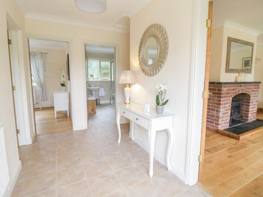 A hallway with tiled floor a white console table with a lamp and plant a round mirror and doors leading to a bathroom bedroom and a room with a brick fireplace at Hendre in Penybontfawr