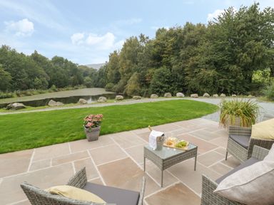 An outdoor patio with wicker chairs and a table with wine and grapes overlooking a grassy area with rocks and a pond surrounded by trees at Hendre in Penybontfawr