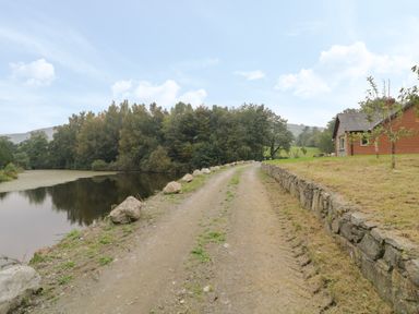 A dirt road beside a pond with trees and a stone wall near a brick house at Hendre in Penybontfawr