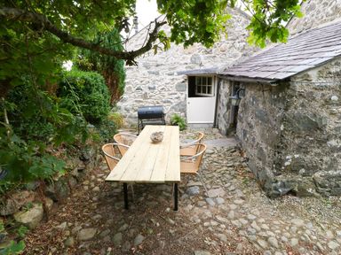 An outdoor patio area with a wooden table and chairs a barbecue grill and stone building walls at Nant Bach Boduan near Nefyn