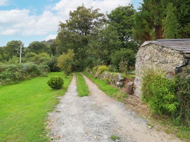 A gravel driveway with grass strips leading past a stone building and trees at Nant Bach in Boduan near Nefyn