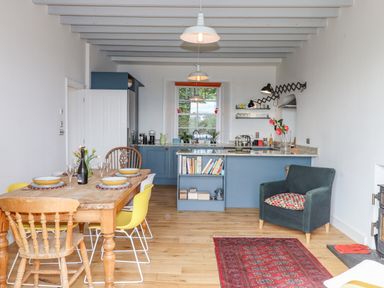 A kitchen and dining area with a wooden dining table set with bowls and glasses yellow chairs blue kitchen cabinets and a dark armchair with a patterned cushion at Wottonsbury in Ilsington