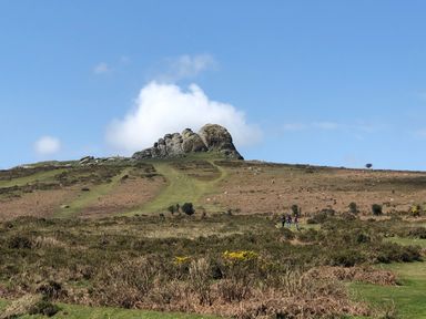 A hill with a rock formation and three people walking near it in a grassy field at Wottonsbury in Ilsington