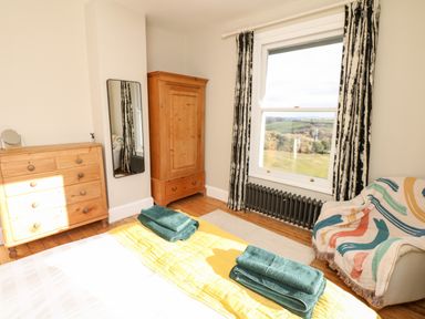 A bedroom with wooden furniture a window with curtains a small armchair with a colorful throw and towels on a bed at Wottonsbury in Ilsington