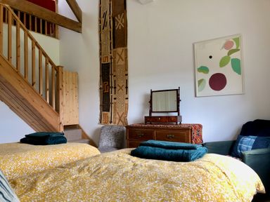 A bedroom with two yellow beds with teal towels a wooden dresser with mirror a patterned chair a staircase and wall art at Wottonsbury in Ilsington