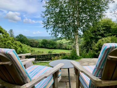 Two wooden chairs with striped cushions and a round table on a patio overlooking green fields and trees at Wottonsbury in Ilsington