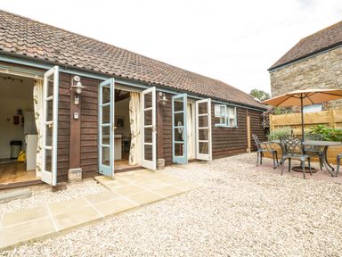 A courtyard with open blue and white doors leading to a wooden building and an outdoor table with chairs and an umbrella at Toms Barn in Mudford Sock near Yeovil Somerset