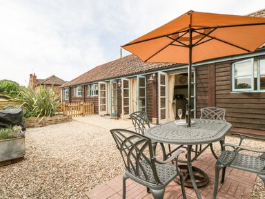 An outdoor patio area with a metal table and chairs under an umbrella in front of a wooden building with open double doors at Toms Barn Mudford Sock near Yeovil Somerset