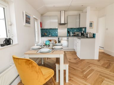 A kitchen with white cabinets a blue tile backsplash a dining table set for six with a yellow chair and wooden bench at Harbour Hideaway in Looe