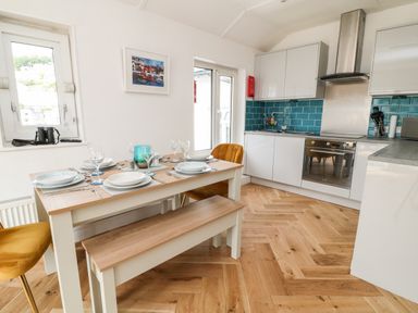 A kitchen and dining area with a wooden table set with plates glasses and cutlery alongside white cabinets and blue tiled backsplash at Harbour Hideaway in Looe
