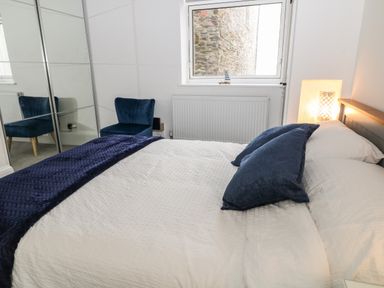 A bedroom with a double bed with blue cushions and blanket a blue chair mirrored wardrobe a radiator and a window at Harbour Hideaway in Looe