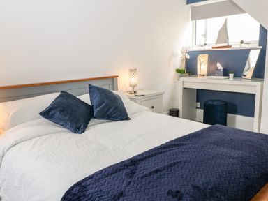 A bedroom with a bed, bedside table with a lamp, a desk with a mirror and lamp, and blue decor at Harbour Hideaway in Looe